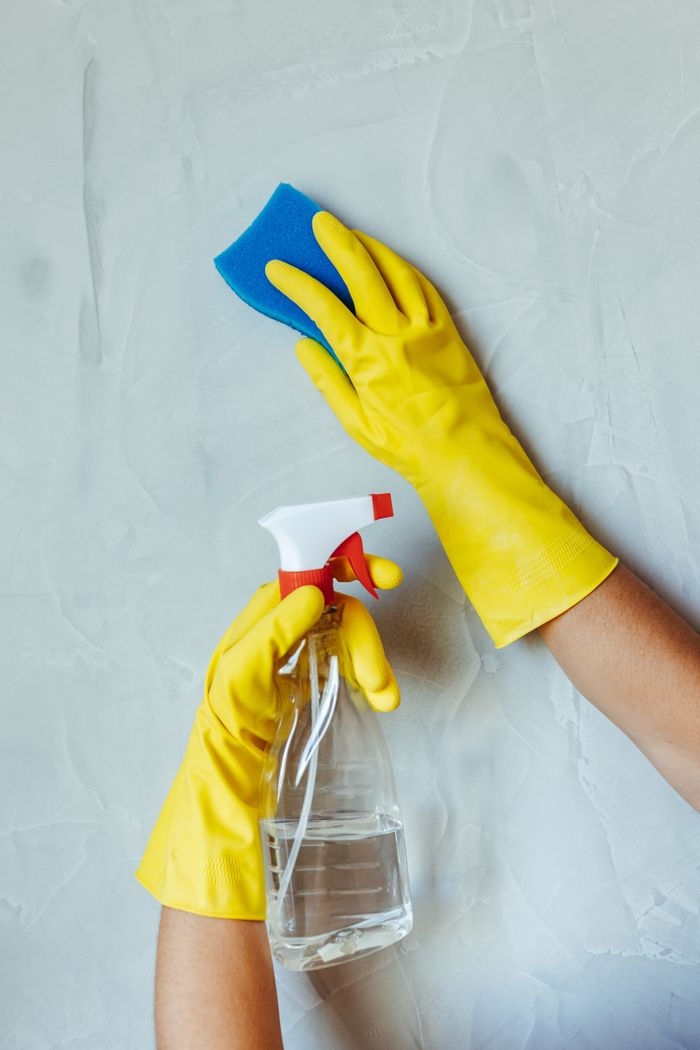 Des mains gantées de jaune nettoient un mur avec une éponge et un flacon pulvérisateur.